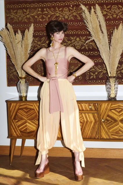Brunette fashion model poses in pink top and sheer beige pants on a wooden floor near a wooden console table with vases.