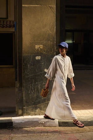 Louise de Chevigny walks in a long white shirt dress and blue baseball cap along a sunlit street beside a stone pillar.