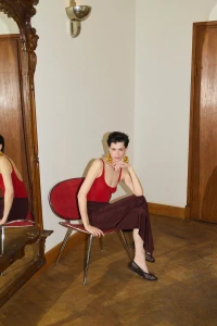 Model seated on a red chair wearing a red tank top and dark trousers in an interior room with a large mirror.