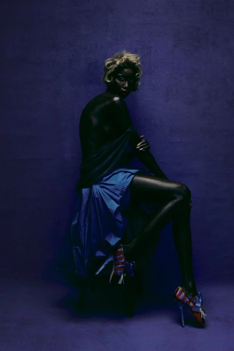 Model with blonde hair and dark skin seated against a purple textured backdrop wearing a blue skirt and patterned heels