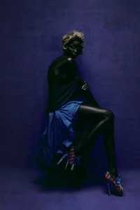 Model with blonde hair and dark skin seated against a purple textured backdrop wearing a blue skirt and patterned heels