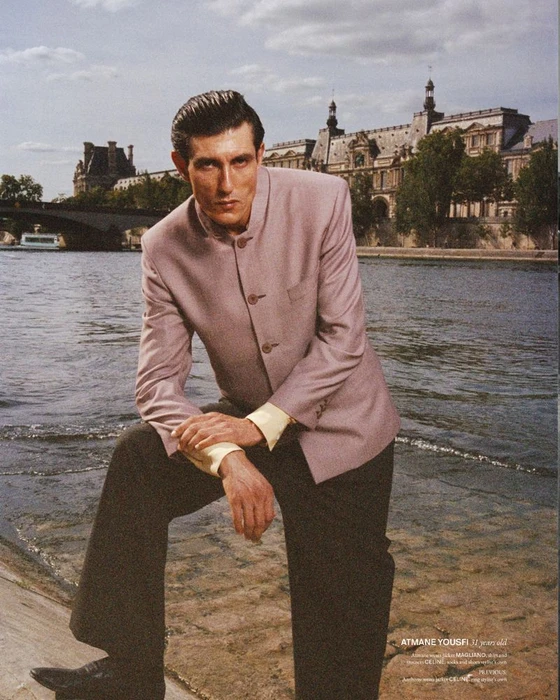 Man wearing a mauve jacket and dark trousers leaning against a stone wall by a river with historic architecture.