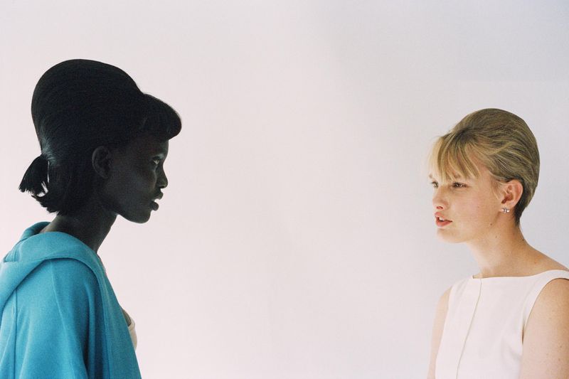 Two women face each other against a plain white background, showcasing 1960s hairstyles and fashion in a profile shot. 