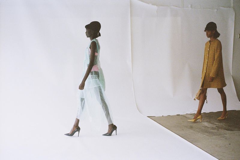 Two African-American fashion models stride confidently against a stark white backdrop, showcasing high heels, sheer dresses, and stylish hats.