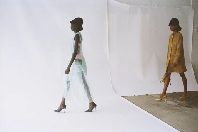 Two African-American fashion models stride confidently against a stark white backdrop, showcasing high heels, sheer dresses, and stylish hats.