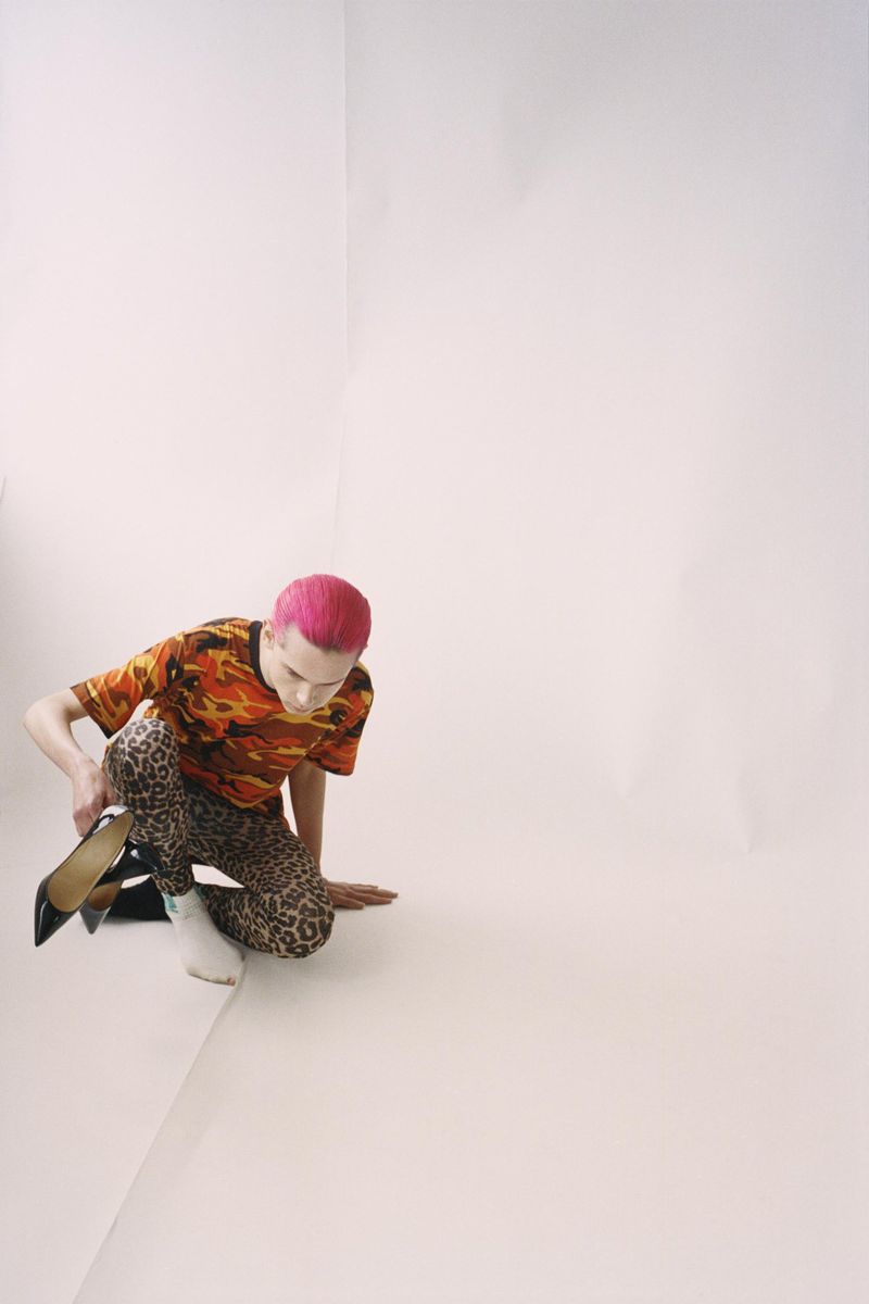 Person with pink hair, orange flame-patterned shirt, and leopard-print leggings squats, holding black high heels in an indoor setting.