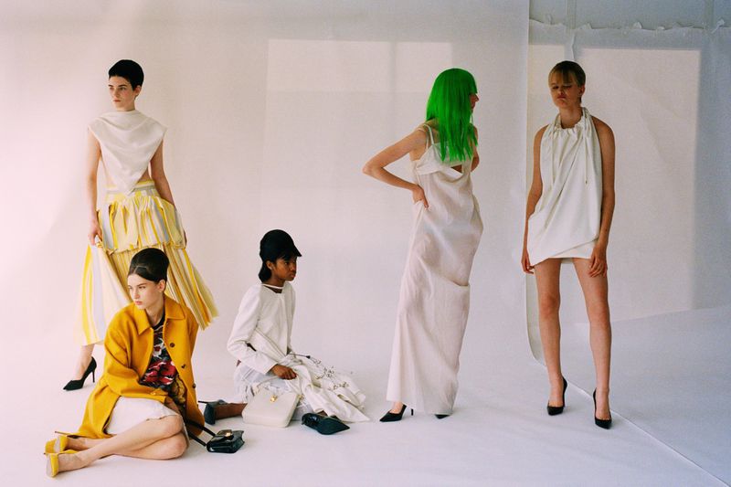 Five female models in avant-garde white and yellow outfits with one model sporting bright green hair in a white studio setting.