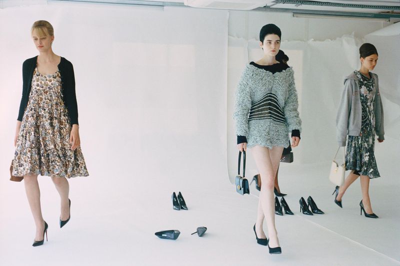 Three fashion models showcase clothing and handbags in a studio setting with scattered black high-heeled shoes on a white floor.