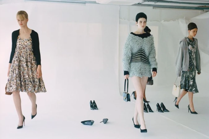 Three fashion models showcase clothing and handbags in a studio setting with scattered black high-heeled shoes on a white floor.