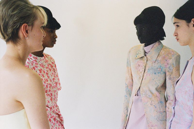 Four diverse women in retro-inspired fashion, facing each other against a plain backdrop, showcasing floral and pastel outfits.