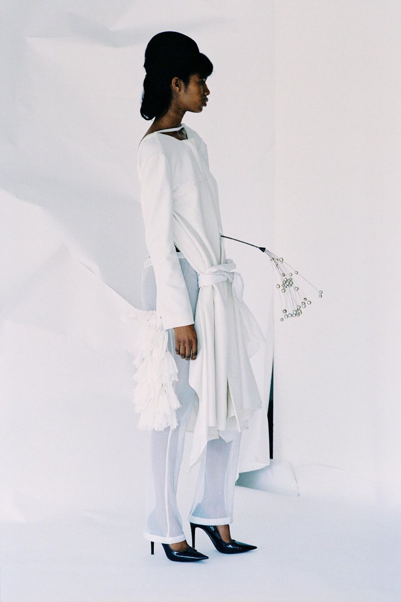 Side profile of an adult woman with dark hair in white top and sheer pants with black heels, holding a small plant on a white background.
