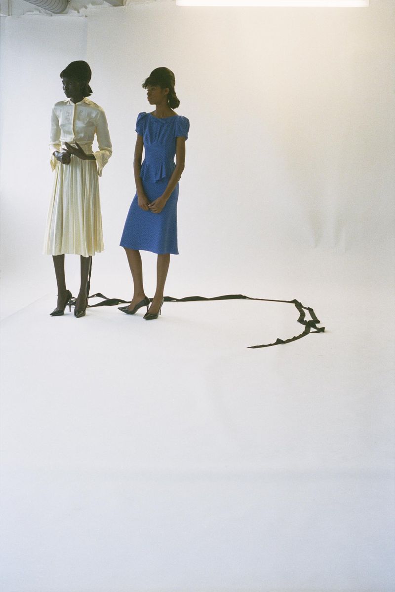 Two women in retro-style dresses and high heels pose in a bright photo studio, featuring a dark trailing ribbon accent.