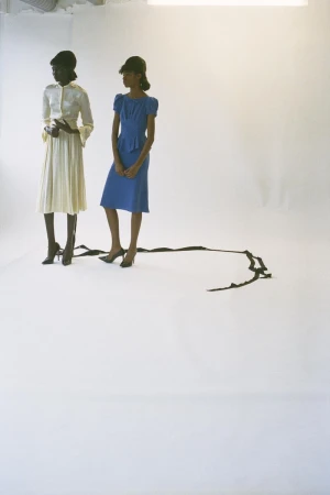 Two women in retro-style dresses and high heels pose in a bright photo studio, featuring a dark trailing ribbon accent.