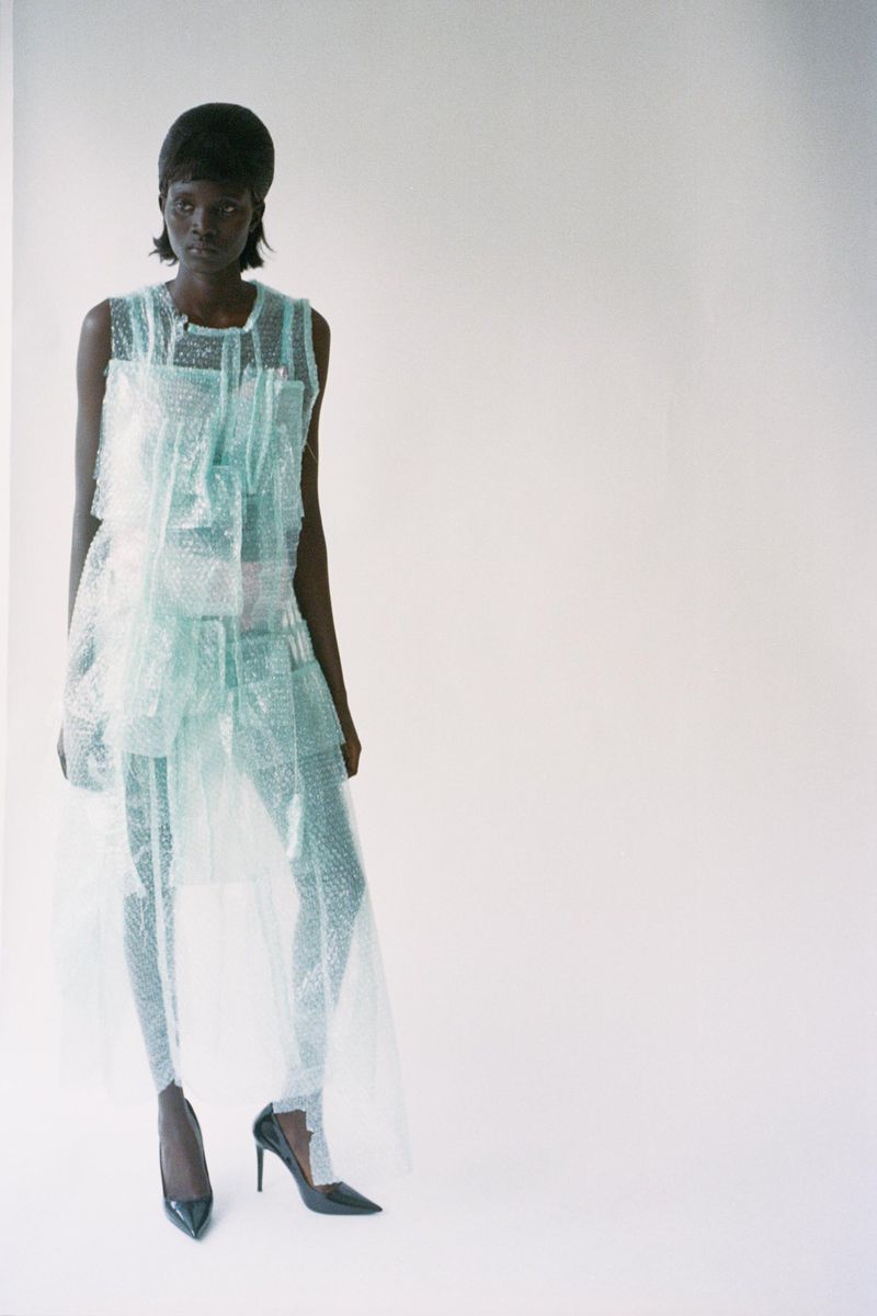 Fashionable dark-skinned model in translucent teal outfit and black patent heels poses in a brightly lit studio with a clean, white backdrop.
