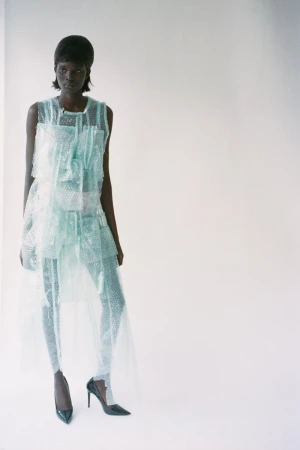 Fashionable dark-skinned model in translucent teal outfit and black patent heels poses in a brightly lit studio with a clean, white backdrop.