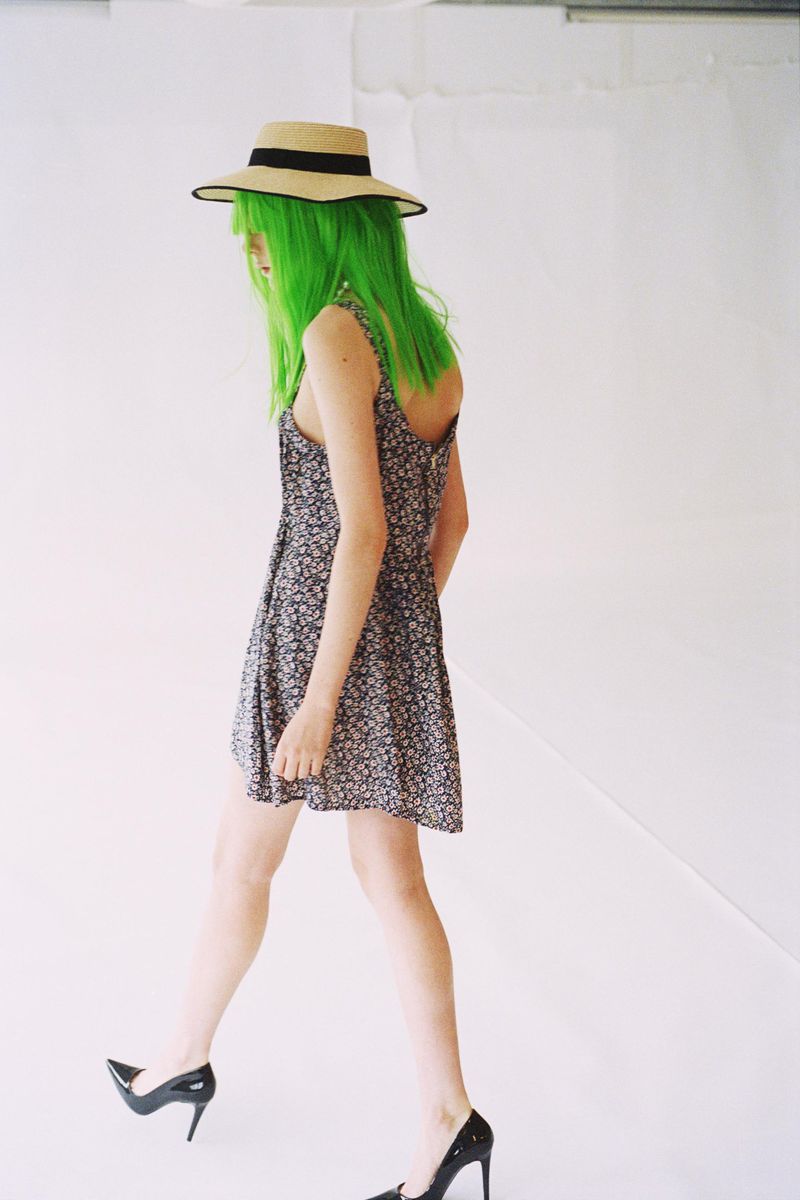 Adult woman in sundress and high heels, wearing a straw hat and green hair, walking on a white seamless background in a studio.
