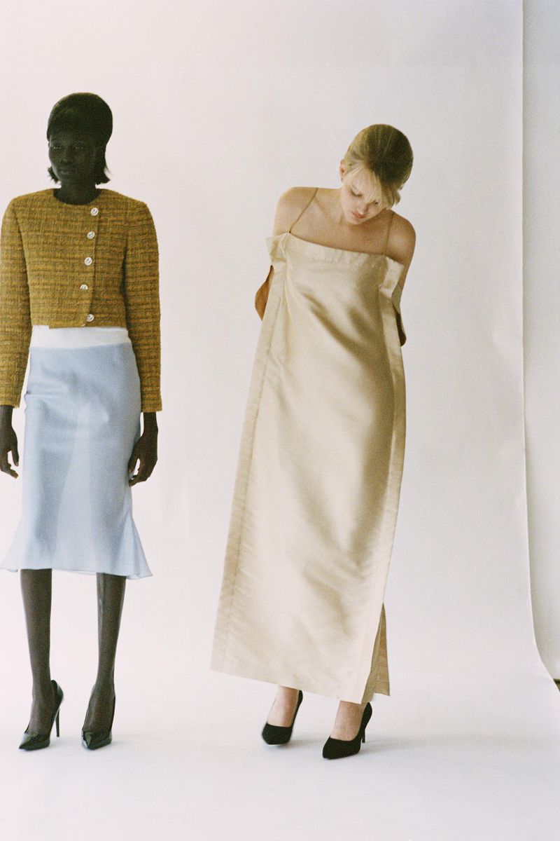 Two women showcasing neutral-toned jackets and dresses in a studio setting, paired with classic black heels, and set against a bright white backdrop.