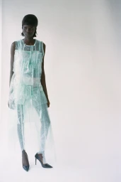 Model in a sheer pale mint textured dress and black pointed-toe stiletto heels standing against a white studio backdrop.