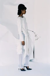 Model in profile wearing a draped white tunic, sheer trousers, and black heels against a crinkled white backdrop.