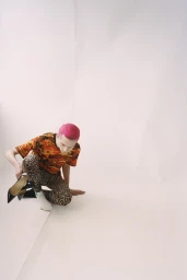 Model with pink hair wearing an orange camouflage t-shirt and leopard print trousers holding heels on white seamless background
