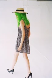 Model with bright green hair wearing a straw hat, floral mini dress, and black heels walking against a white studio background.