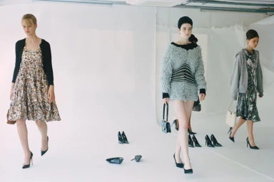 Three models walk across a white studio floor scattered with black high-heeled pumps wearing eclectic knitwear and dresses.