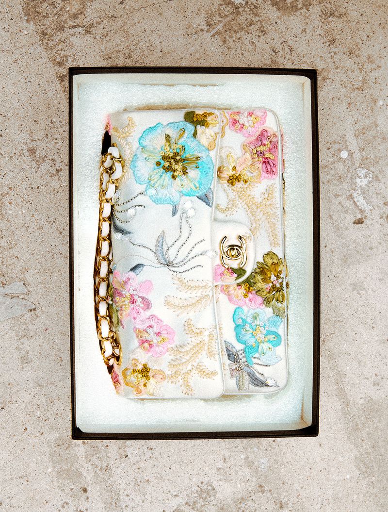 White embroidered handbag with colorful floral beadwork and gold chain in a presentation box on a textured surface.