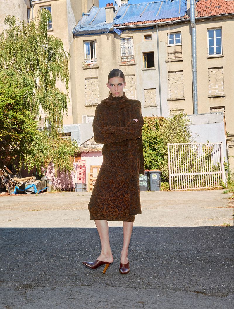 Model standing outdoors wearing a brown patterned outfit with a matching scarf and dark heels in front of an apartment building