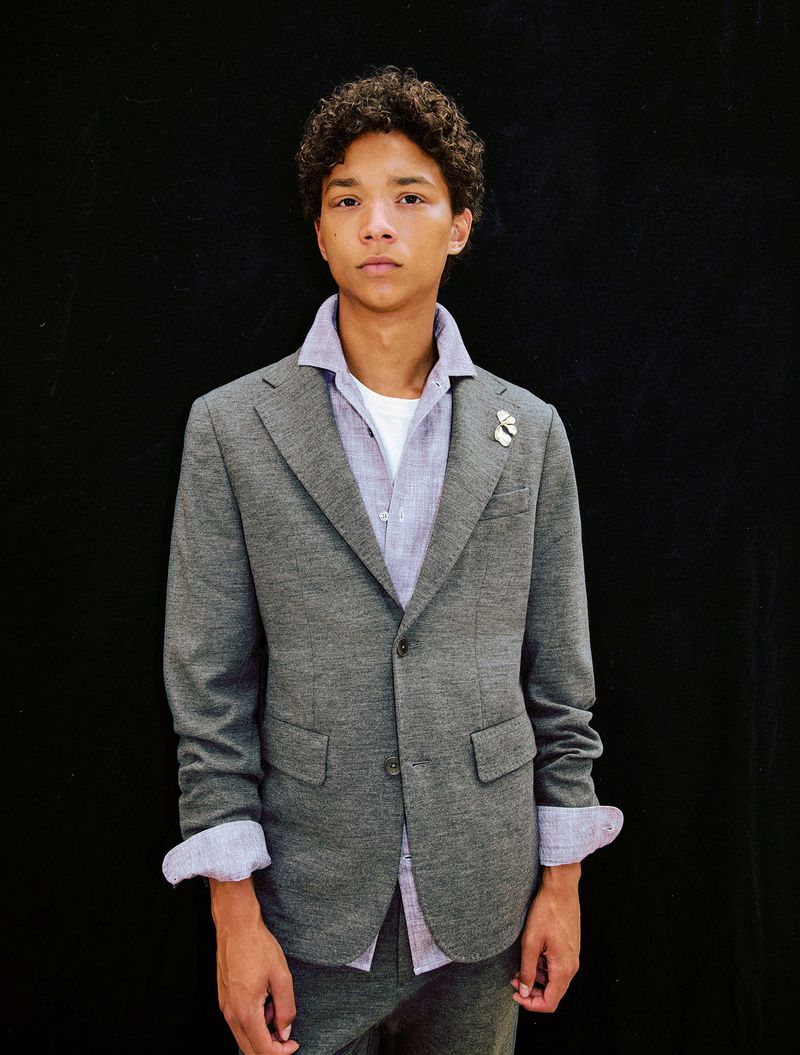 Portrait of a young male model with curly brown hair wearing a grey blazer over a purple shirt against a dark backdrop