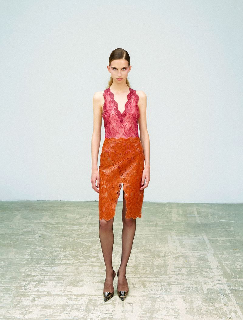 Model standing on a worn concrete floor against a plain white wall wearing a pink lace top and orange lace skirt.