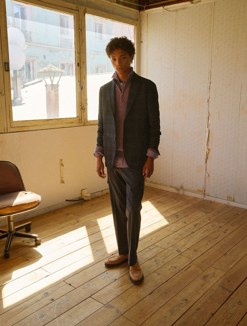 Young male model standing in an empty room with a wooden floor wearing a dark suit jacket, purple sweater, and trousers.