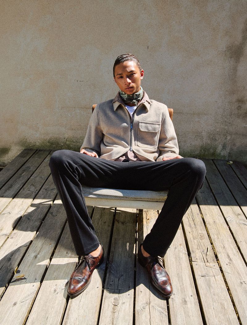 Model seated on a chair on a wooden deck wearing a textured jacket, black trousers, and patterned neck scarf against a textured wall.