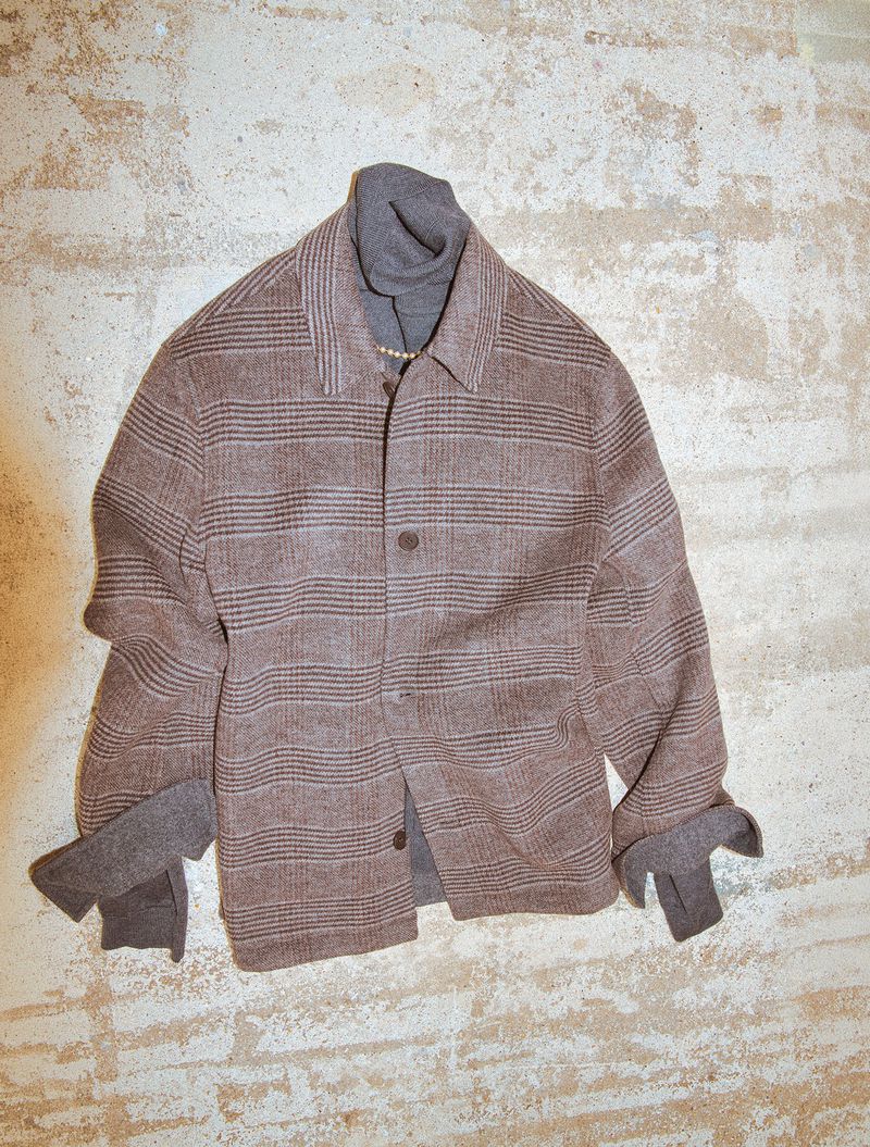 Brown plaid jacket layered over a grey turtleneck against a textured stone wall background