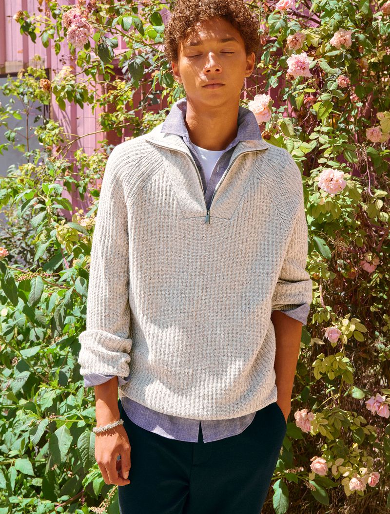 Young man with curly brown hair wearing a cream quarter-zip ribbed sweater standing outdoors before rose bushes.