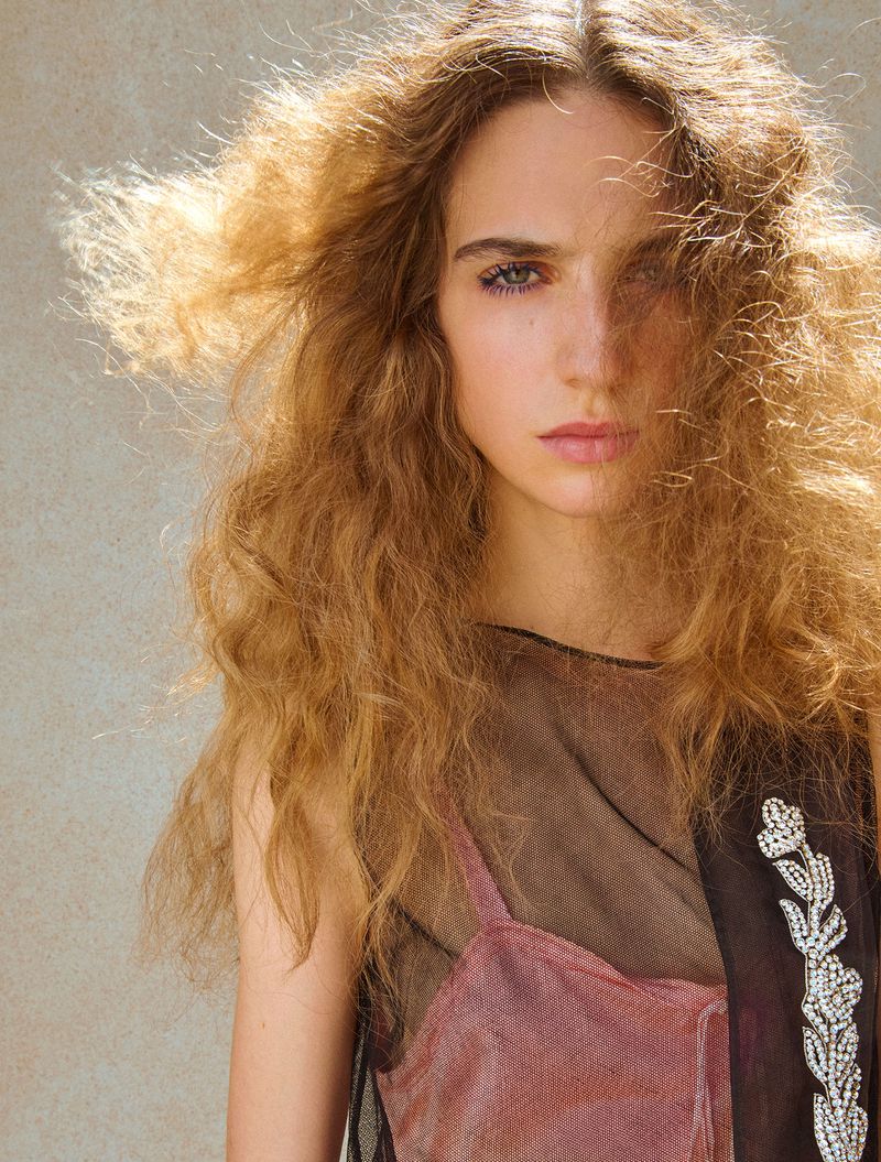 Close-up portrait of a woman with voluminous blonde curly hair wearing a pink slip dress and dark sheer mesh overlay.