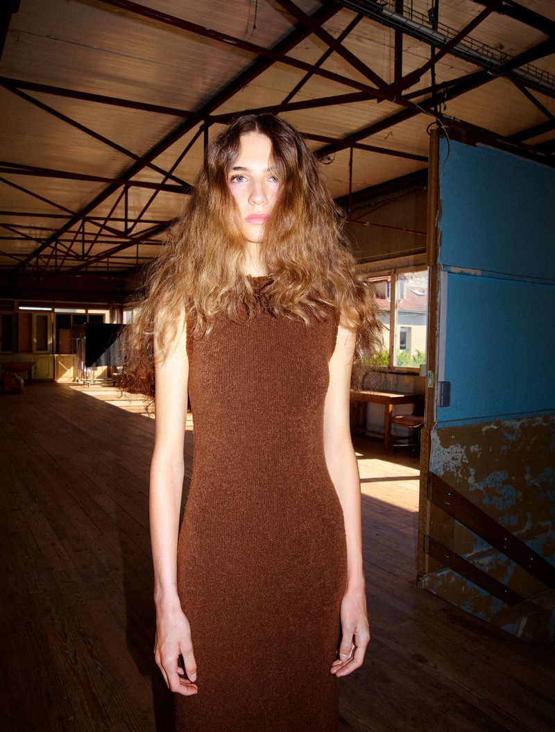 Model with long wavy brown hair wearing a sleeveless brown knit dress in a rustic wooden interior for Family Style.