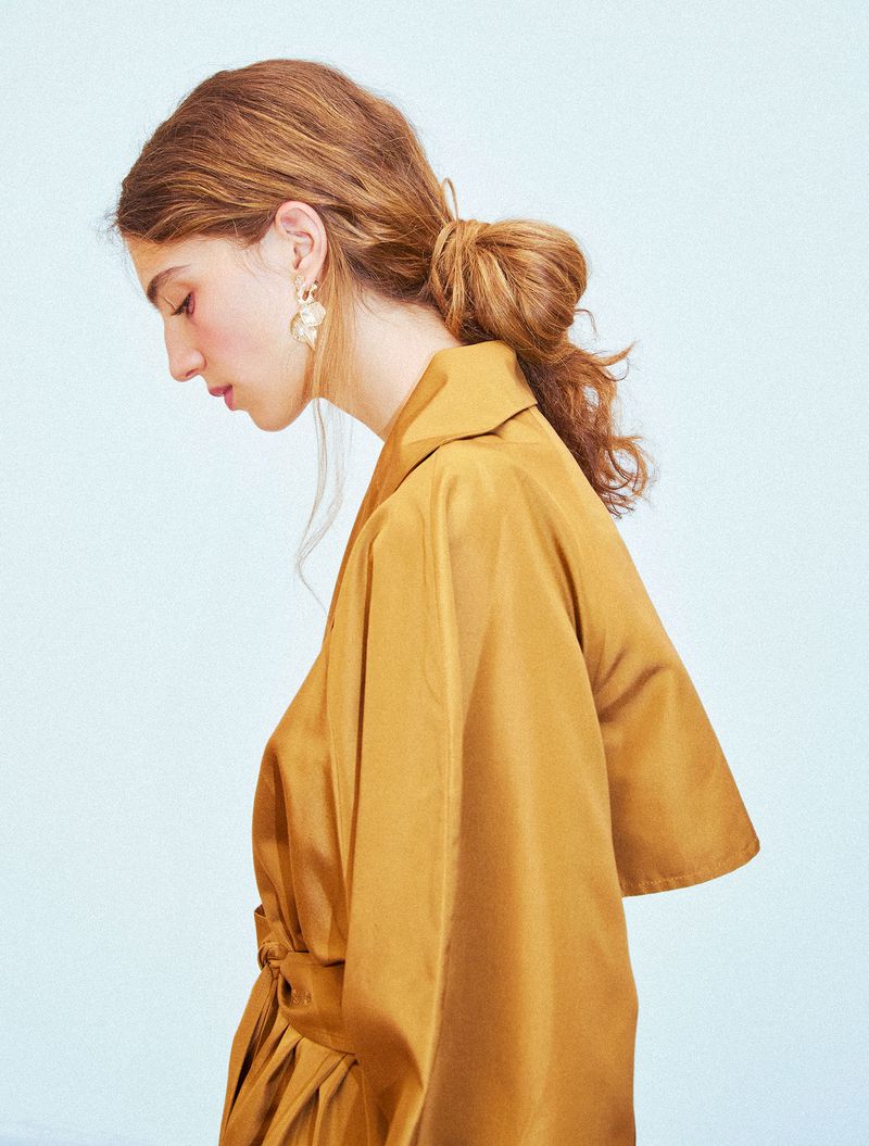 Profile portrait of a model with reddish-brown hair tied in a loose low bun wearing a mustard yellow silk garment and ornate gold earring