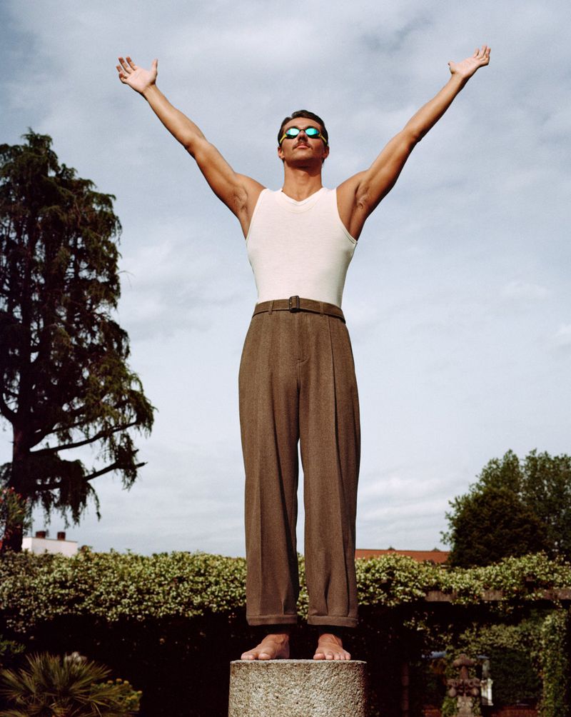 Adult male stands on stone plinth with arms raised, wearing sunglasses, tank top, and high-waisted trousers in an outdoor setting. 