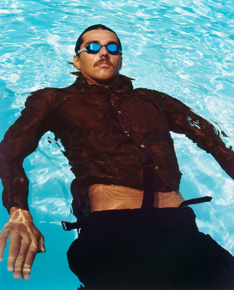 Man in a brown shirt and black pants swims in a pool while wearing blue goggles. Water reflects light.