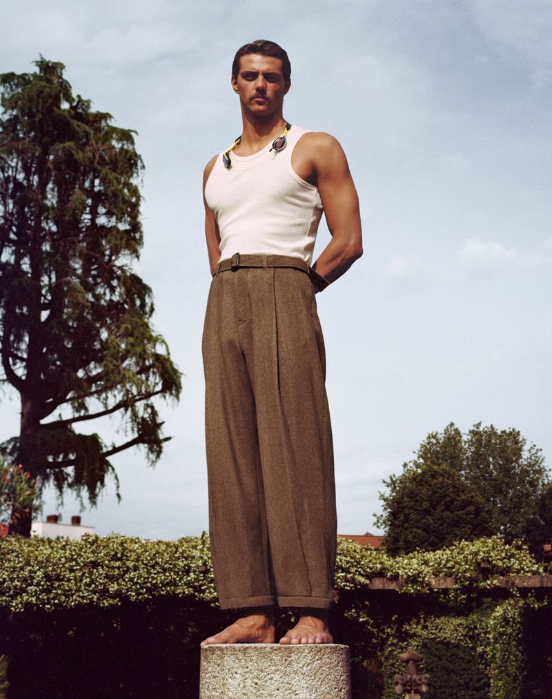 Male fashion model stands barefoot in wide-legged trousers and tank top on stone plinth outdoors in a formal garden setting under sky.