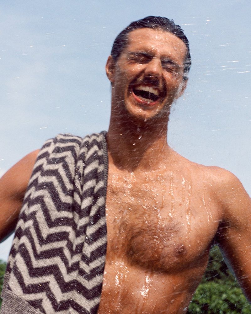 Shirtless man laughs with water cascading down, wearing a zig-zag pattern towel over his shoulder against a light blue sky.
