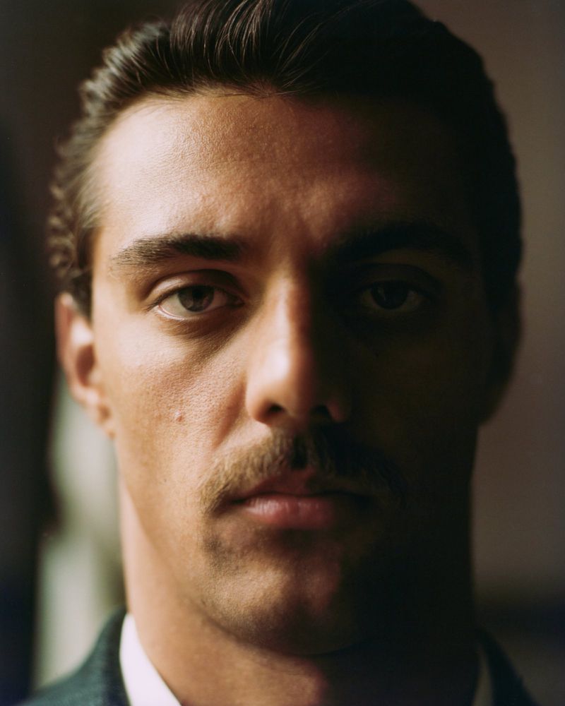 Close-up portrait of a man with dark hair and a mustache looking directly at the camera, wearing a suit jacket in a dimly lit setting.