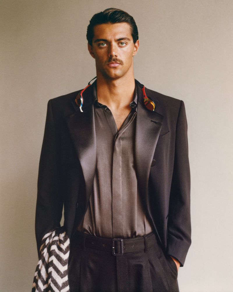 Fashionable adult man in a dark suit and grey shirt with a patterned shawl, posing in an indoor studio setting, close-up shot.