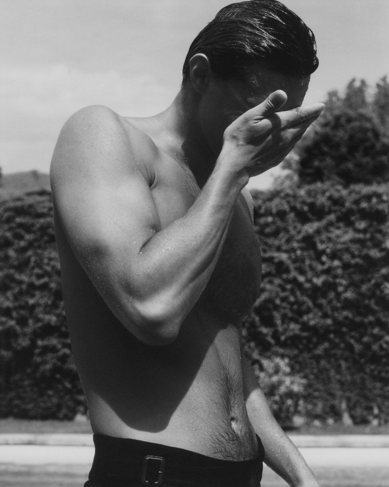 Monochrome image of a shirtless adult male in front of greenery, covering his face with his hand in an outdoor setting.