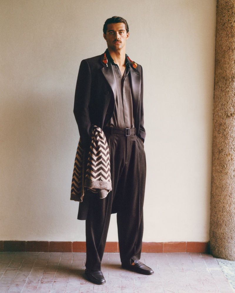 A full-length portrait showcases a man in a dark suit with a patterned jacket draped over his arm, set against a neutral interior wall.