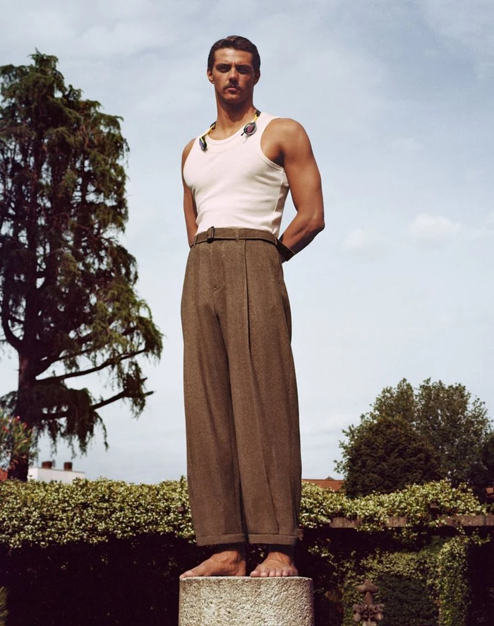 Man wearing a white tank top and brown trousers standing outdoors with trees in the background