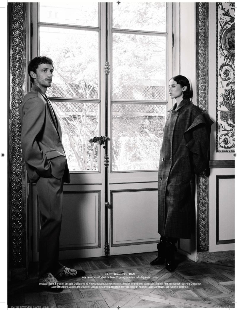 Black and white editorial portrait of a man in a suit and a woman in a long coat standing before tall French doors.