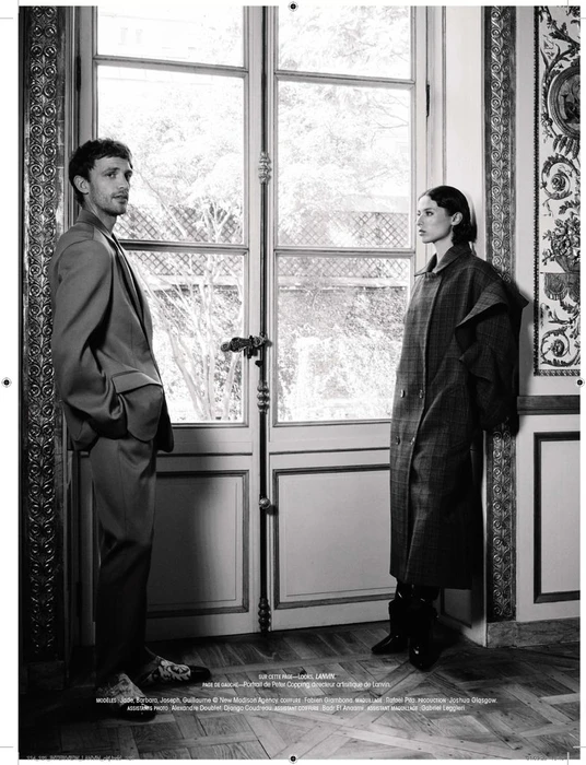 Black and white editorial portrait of a man in a suit and a woman in a long coat standing before tall French doors.