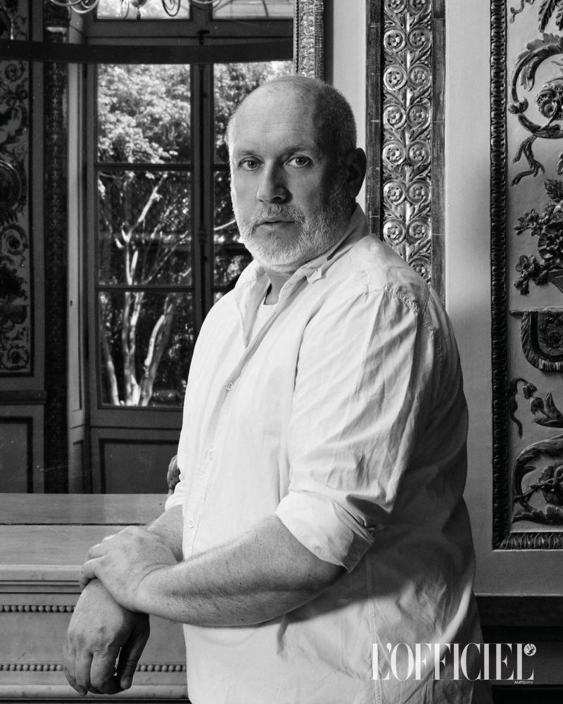 Portrait of Peter Copping wearing a white button-down shirt leaning against a mantelpiece in an interior setting.