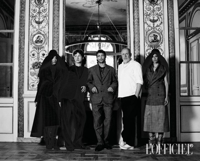 Monochrome fashion portrait featuring five individuals posing in an ornate room with classical wall panels and a large chandelier.